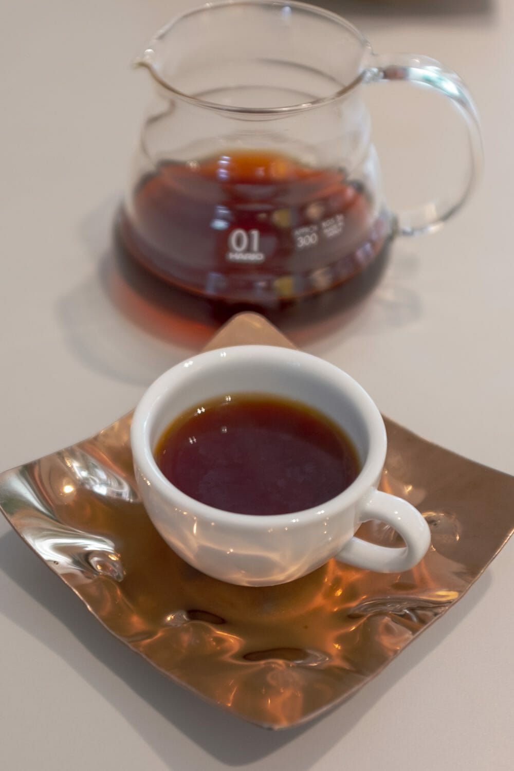 Coffee mug full of coffee on a copper tray with a glass serving carafe in the background.