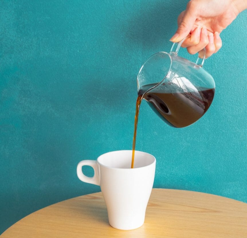 Hand pouring coffee from a glass carafe to a mug with a blue background.