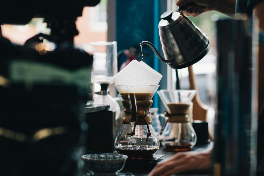 Coffee kettle slowly dripping over a chemex with blurred coffee equipment in the background